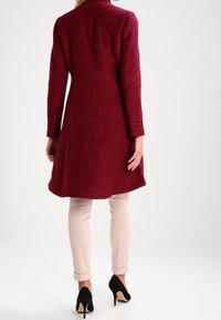 Lang, getailleerde bordeauxkleurige jas van textuurstof, met een hoge kraag en twee zijzakken. Draagt over lichte broek en zwarte hakschoenen.