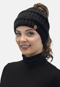 Zwarte gebreide beanie met een textuur ribbelpatroon, featuring een klein bruin leren logo etiket aan de zijkant. Draagt over een zwart turtleneck.