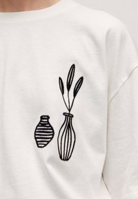 Witte T-shirt met zwarte borduursels van twee vazen en bladeren; de ene vaas heeft horizontale strepen, de andere is omrand met verticale lijnen.