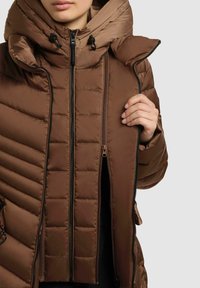 Braune gepolsterte Jacke mit hohem Kragen und Reißverschluss, die kontrastierende gesteppten Strukturen und glatte Stoffabschnitte aufweist.