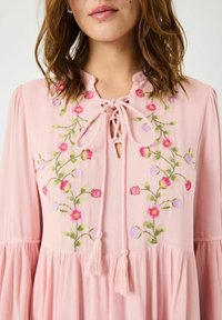 Blusa rosa realizzata in tessuto strutturato, caratterizzata da ricami floreali nei colori rosa, viola e verde, con scollatura a forma di chiave e dettaglio con fiocco.