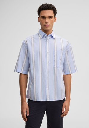 Młody mężczyzna w krótkim, jasnoniebieskim koszula zapinana na guziki z pionowymi białymi i beżowymi paskami oraz ciemnymi spodniami, stojący na jednolitym tle.