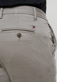 Beiga byxor med bakficka som knäpps, och en liten röd, vit och blå flagglogotyp, kombinerade med en svart topp synlig vid midjan.