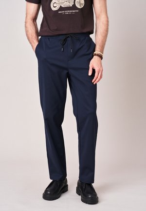 Man draagt marineblauwe broek met trekkoorden, zwarte laarzen en een donkerbruin T-shirt met een motorfietsafbeelding, staat met handen in de zakken.