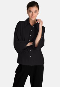 Camicia nera con bottoni, colletto ampio, maniche morbide e bottoni lucidi. Il tessuto ha una consistenza liscia.
