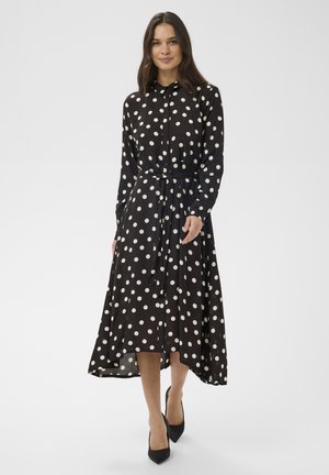 Femme portant une robe chemise midi noire à pois blancs, manches longues, fermeture par boutons et ceinture à la taille, associée à des talons noirs.