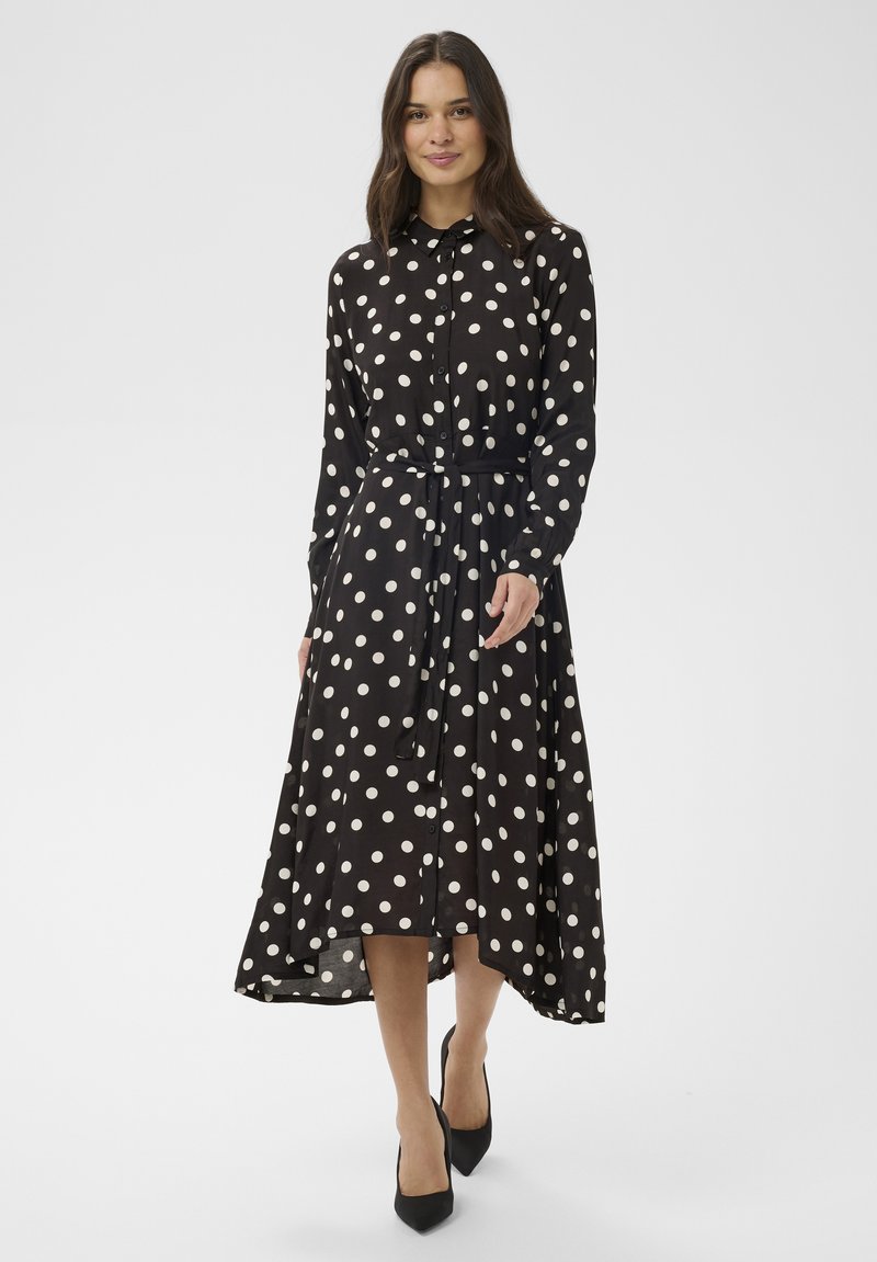 Femme portant une robe chemise midi noire à pois blancs, manches longues, fermeture par boutons et ceinture à la taille, associée à des talons noirs.