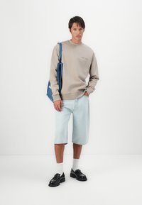 Svetlo šedý mikina, voľné džínsy šortky, čierne mokasíny a biele ponožky. Modrá taška s bielym textom. Jednoduchý, voľný outfit.