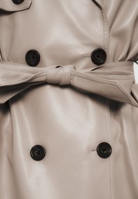 Μπεζ δερμάτινο παλτό τρεντς με ομαλή υφή, με μαύρα κουμπιά, ζώνη στη μέση και κλασικό γιακά.