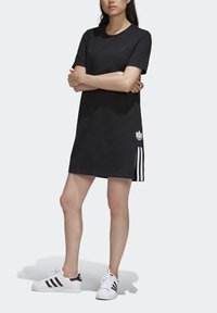 Kvinna står med armarna i kors iförd en svart Adidas kortärmad klänning och vita Adidas sneakers med svarta ränder.