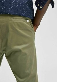 Olive green pants with a smooth texture and tapered design, featuring side pockets and a small white brand label on the rear.
