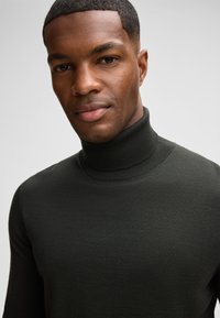 Nahaufnahme eines Mannes mit einem dunkelgrünen Rollkragenpullover, der einen selbstbewussten Ausdruck und eine glatte, klare Haut zeigt. Der Hintergrund ist neutral grau.