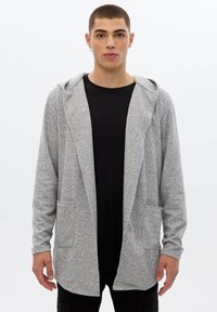 Joven con cabello corto, llevando un cárdigan de punto gris abierto con bolsillos sobre una camiseta negra, de pie frente a un fondo blanco.