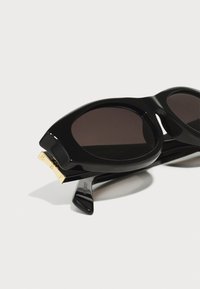 Occhiali da sole oversize neri con lenti scure e montatura in plastica lucida. Presentano un accento dorato sulla tempia con testo inciso.