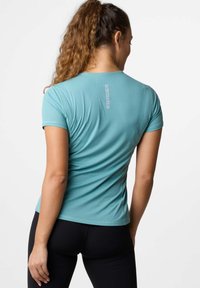 Maglietta sportiva turchese a maniche corte, in tessuto strutturato, con un dettaglio del logo sulla parte superiore della schiena. Abbinata a pantaloni sportivi neri.