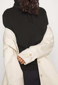 Černá mikina s kapucí a šnůrkami, položená pod bílým, oversize kabátem s knoflíkovou manžetou. Hladký materiál s kontrastními barvami.