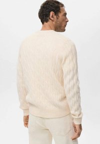 Kremowy sweter w grubym splotem warkoczowym z prążkowanymi mankietami i dołem, z teksturowanym wzorem, noszony z jasnymi spodniami.