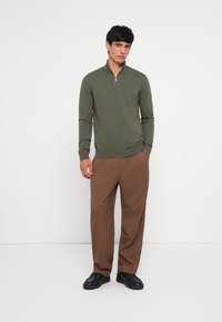 Olive green zip-neck sweater made of soft fabric, paired with brown wide-leg trousers and black sneakers. Simple design, no visible patterns.