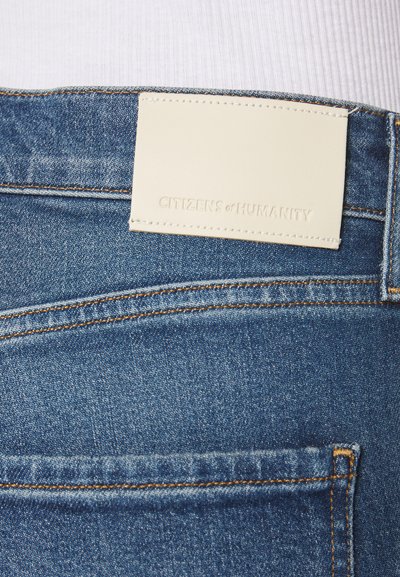 Jeans in denim blu scuro con una toppa in pelle di colore chiaro che presenta testo impresso, cuciture rinforzate e un design standard delle tasche.
