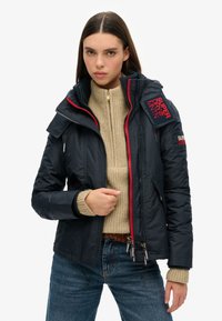 Veste matelassée bleu marine avec des accents de fermeture éclair rouge, col haut, plusieurs poches zippées et un pull côtelé beige clair en dessous.