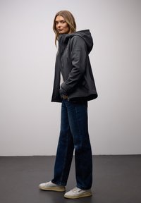 Femme portant une veste à capuche sombre, un jean bleu et des baskets blanches, debout de profil avec les mains dans les poches devant un fond uni.