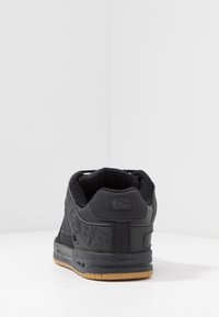 Sneaker nera con suola testurizzata e collare imbottito vista da dietro su una superficie bianca contro un muro angolare grigio chiaro.