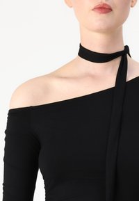 Juodas of-the-shoulder viršus, pagamintas iš lengvos medžiagos, turintis siaurą raištį aplink kaklą ir lygią tekstūrą. Be raštų ar akcentų.
