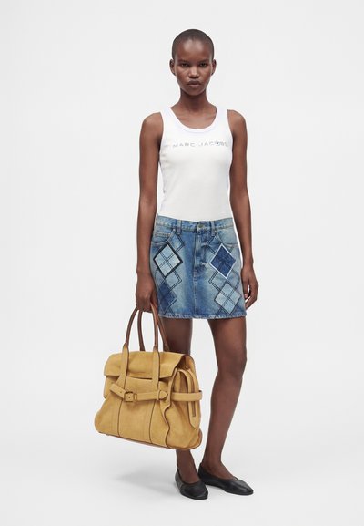 Femme portant un top blanc sans manches et une jupe en jean bleue avec des patchs en forme de diamant tient un grand sac à main en cuir marron clair et porte des ballerines noires.