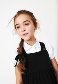 Czarna sukienka z guzikami na ramionach, warstwową spódnicą i białą koszulą z kołnierzykiem. Brązowe warkocze ozdobione czarną kokardą z lewej strony.