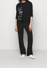Persona che indossa un felpa nera con testo bianco multilingue e pantaloni neri con testo laterale, abbinati a sneakers bianche, su uno sfondo semplice.