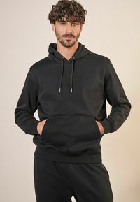 Svart huvtröja i mjukt material, med en framficka, dragsko i huvan och ribbad mudd vid ärmarna och nederkanten.
