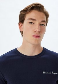 Navy blue ribattipaita, joka on valmistettu puuvillasta, pyöreällä kaula-aukolla ja valkoisella brodeeratulla tekstillä "Unelmoi inspiroidaksesi" rintakehässä.