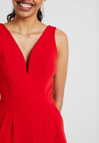 Vestido rojo sin mangas con un escote en V profundo, cintura ajustada y falda plisada. La tela suave y las líneas limpias crean una silueta elegante.