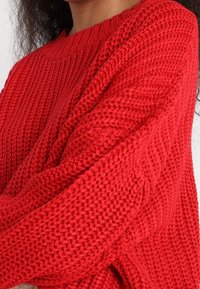 Maglione rosso lavorato a maglia con una texture spessa, dotato di maniche lunghe e scollo rotondo. Il tessuto presenta un motivo a coste.