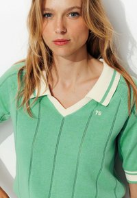 Pull vert avec un col crème, présentant des rayures verticales et un emblème '75' sur la poitrine. Tissu doux et texturé.