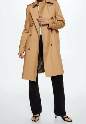 Personne portant un trench-coat beige croisé, un pantalon noir et des sandales noires à talons ouvertes, debout devant un fond uni.