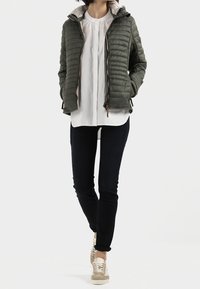 Olijfgroene pufferjack met een gequilt ontwerp, voorzien van een capuchon en ritsluiting. Draag het over een wit shirt, in combinatie met donkere spijkerbroeken en beige sneakers.