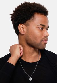 Zilveren ketting met een ronde hanger met ingewikkelde ontwerpen, gedragen door een persoon met kort, krullend haar, gekleed in een zwart shirt.