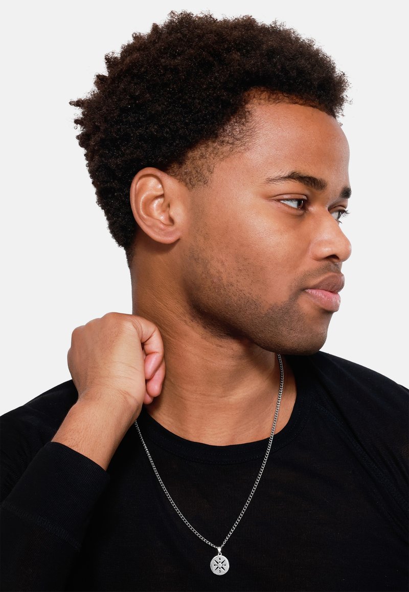 Zilveren ketting met een ronde hanger met ingewikkelde ontwerpen, gedragen door een persoon met kort, krullend haar, gekleed in een zwart shirt.