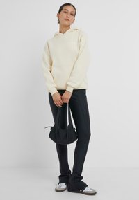 Krämfärgad hoodie med framficka och dragsko i huvan, kombinerad med svarta slim-fit byxor och en liten svart axelväska. Sneakers med vita detaljer.