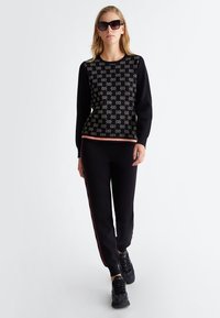 Maglione nero con texture stampata, bordi rossi e pantaloni neri abbinati con strisce laterali. Modello che indossa occhiali da sole e sneakers.