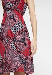 Rotes Kleid mit einem Paisley-Muster in Rosa und Schwarz, ausgestattet mit einem taillierten Oberteil, einem ausgestellten Rock und einer gebundenen Taille. Glatte Stofftextur.
