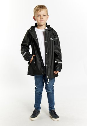 Sort vandtæt regnjakke med hætte, reflekterende detaljer, trykknapper og en frontlomme, båret over en hvid skjorte og blå jeans.