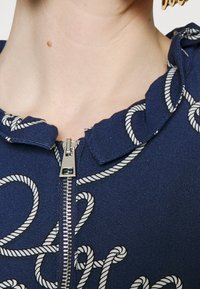 Mörkblå zip-up plagg med en volangkrage och vit snöre-liknande broderi. Dragkedjan är metallisk och placerad centralt.