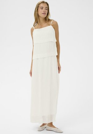 Femme portant une robe maxi blanche plissée à plusieurs couches avec des bretelles fines et des chaussures plates scintillantes, posant devant un fond uni.