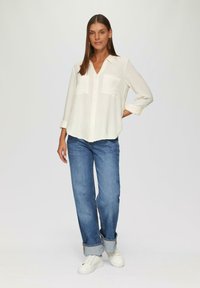 Weißes, langärmeliges Hemd mit Knöpfen und zwei Brusttaschen, kombiniert mit hellblauen, geraden Jeans und weißen Sneakern.