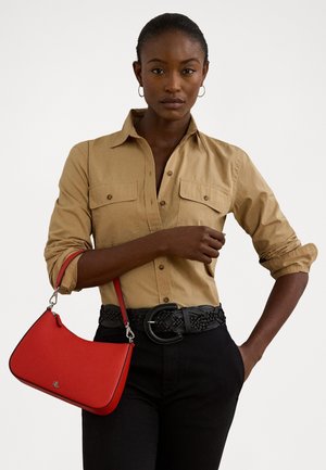 Femme portant une chemise beige boutonnée et un pantalon noir, tenant un petit sac à main rouge sur son bras, sur un fond uni.