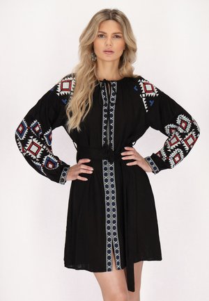 Zwarte jurk met lange mouwen, voorzien van ingewikkelde rode, witte en blauwe geometrische borduursels, een gestrikt taille en een voorste split.
