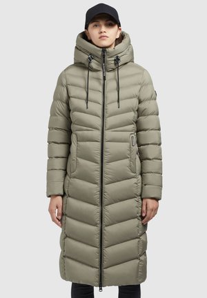 Femme portant un long manteau matelassé vert olive avec un col haut, une fermeture éclair noire et des cordons, associé à une casquette noire et un pantalon noir.
