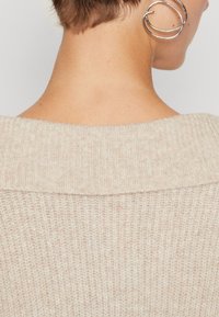 Beige stickad tröja med en bred, vikbar krage, som har en mjuk textur och ribbat mönster. Silvrig cirkulär örhänge synlig vid örat.